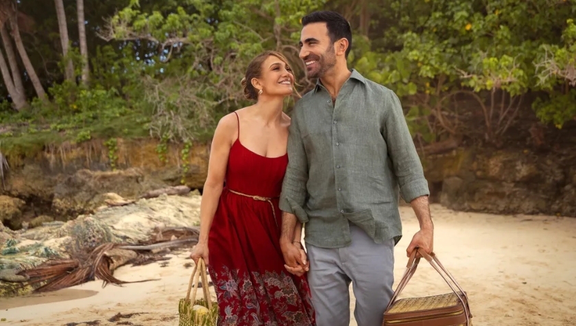 A couple walks hand-in-hand along a tropical beach, smiling at each other, carrying picnic baskets
