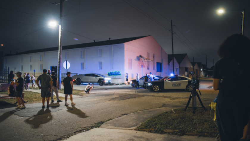 A film crew shoots a nighttime exterior scene on a city street with police cars in frame
