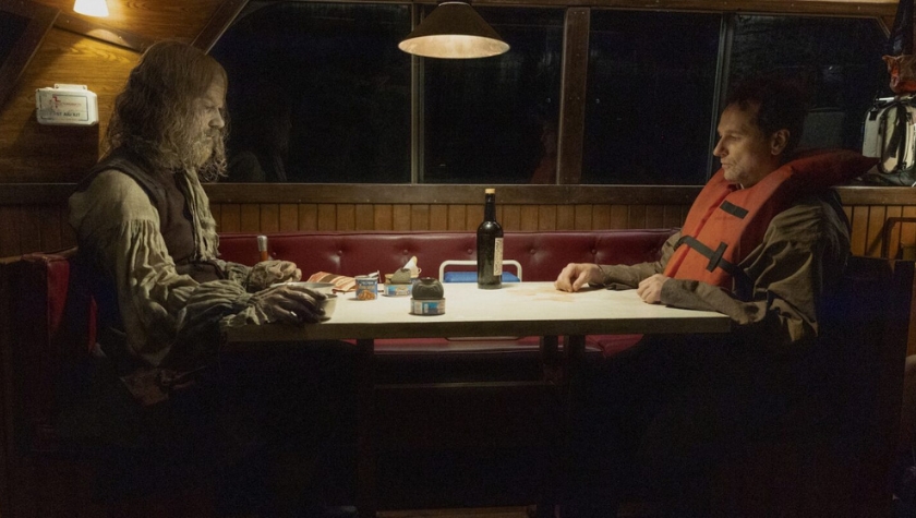 Two men seated across from each other in a dim boat diner — one disheveled with a long white beard, the other in an orange life jacket