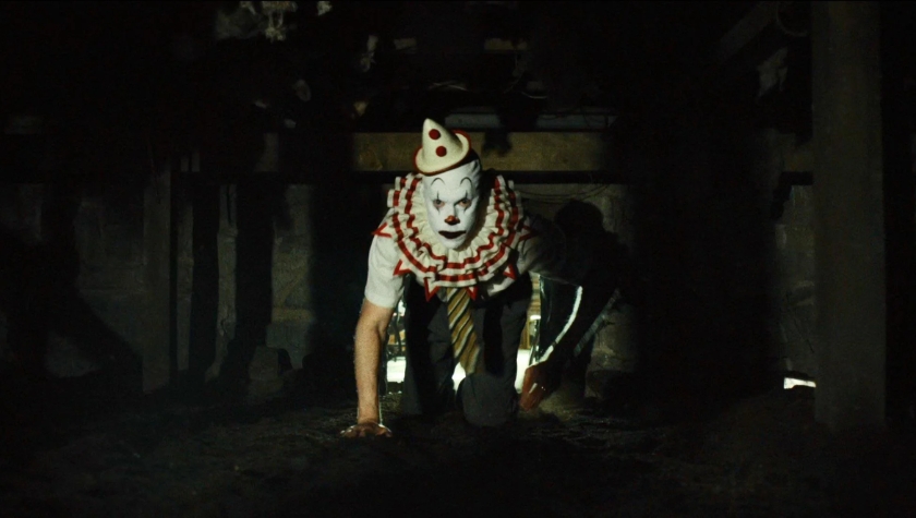 A terrifying clown in white face paint and red accents crawling on all fours through a dark crawlspace