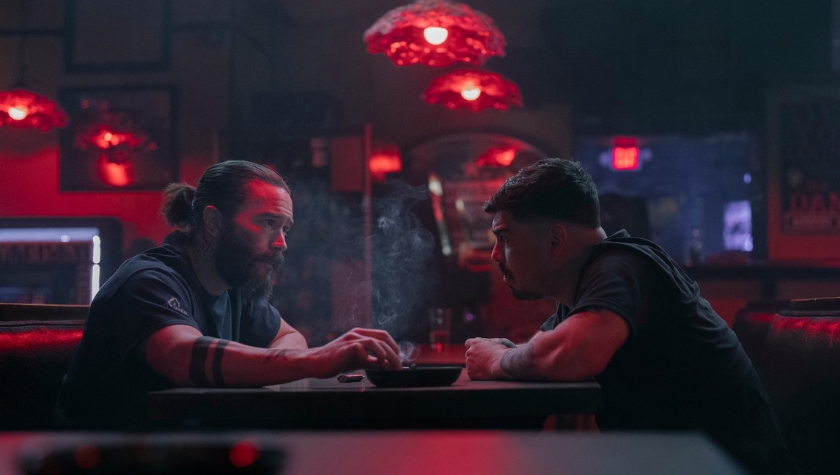 Two men face each other across a table in a dimly red-lit bar in a scene from Task on HBO