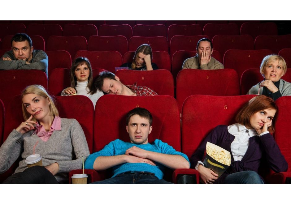 Group of people in a movie theater appearing bored or uninterested, with some yawning and others slouching in red seats.