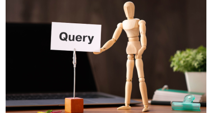 a wooden figurine gesturing towards a white card displaying the word Query. A laptop lays on the desk in the background