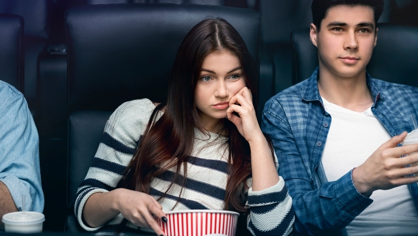 Young couple sitting in a movie theater looking bored, with the woman resting her chin on her hand and holding popcorn.