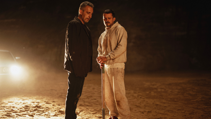 Image of Orlando Bloom and John Turturro looking down at the ground. Orlando Bloom holds a shovel, and a parked car shines a headlight onto the two of them.