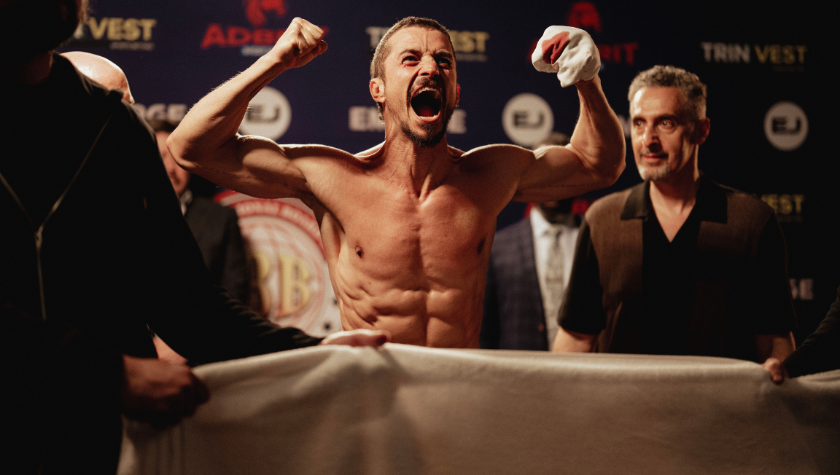 Image of Orlando Bloom jumping up and down cheering with 2 raised firsts, one of which is wrapped in a bloody cloth. John Turturro stands behind smiling
