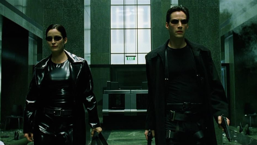 Keanu Reeves and Carrie-Anne Moss, both hands holding guns, walk down a hallway in 'The Matrix'