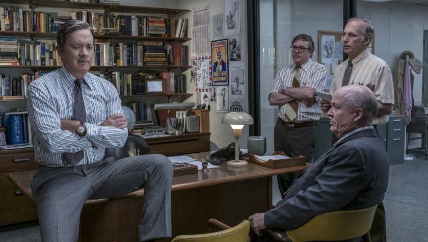 Tom Hanks sits on his desk thinking, and John Rue sitting opposite, as David Cross and Bob Odenkirk lean against a glass office window in The Post