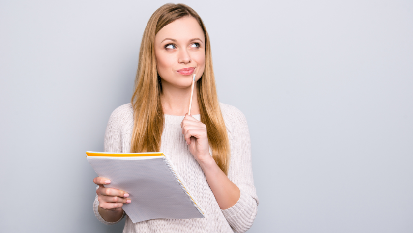 Writer stands with a notepad. She holds a pencil to her face, thinking