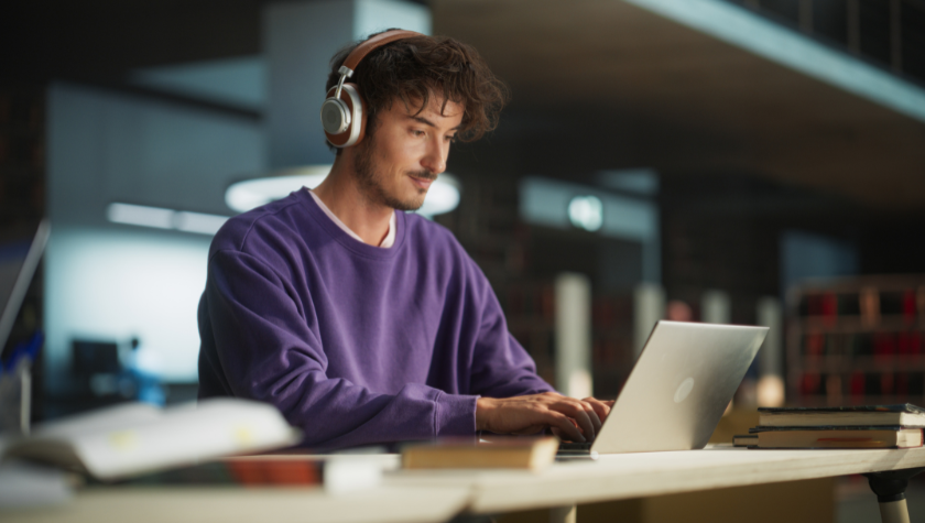 man writing on his laptop in a library wearing headphones